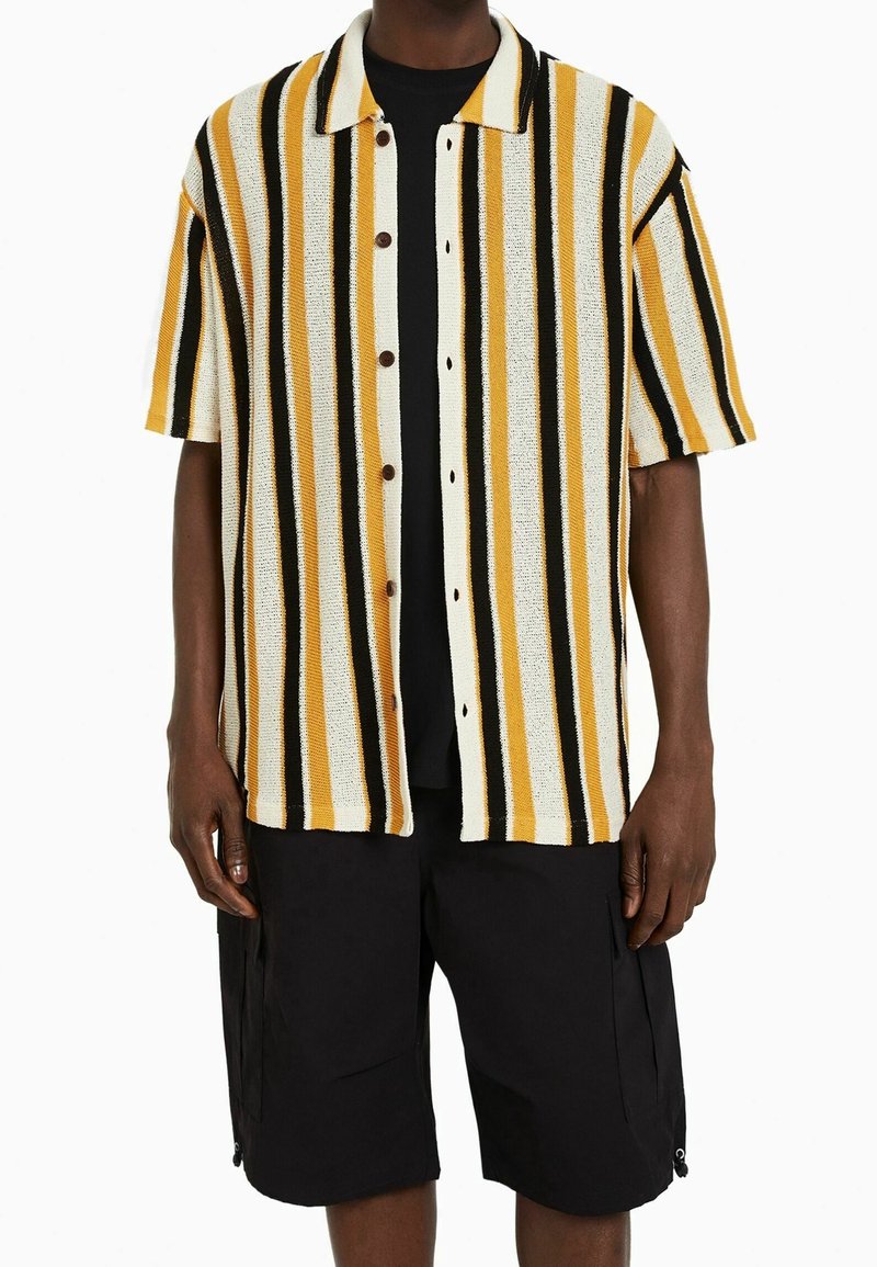 Koszula z krótkim rękawem w pionowe paski w kolorze żółtym, czarnym i białym, z zapięciem na guziki z przodu i kołnierzykiem rozłożonym. Połączona z czarnymi szortami.