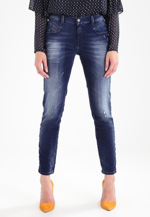 Femme portant un jean skinny bleu foncé usé, un chemisier noir à pois et des chaussures à talons hauts orange vif, debout, mains détendues.