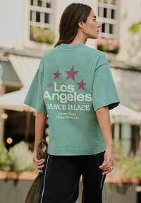 Tricou supradimensionat verde cu stele mov și text alb. Printul de pe spate spune "Los Angeles DANCE PALACE" și detalii despre locație dedesubt.