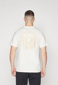 T-shirt en coton blanc avec un grand graphique circulaire beige au dos, incluant les textes « PARIS » et « SAINT-GERMAIN » ainsi qu'une silhouette de joueur de basketball.