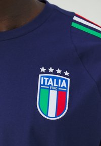 Granatowa koszula z logo "ITALIA" i tarczą, z zielonymi, białymi i czerwonymi paskami, oraz czterema białymi gwiazdami nad emblematem.