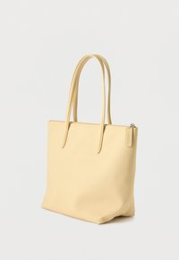 Sac cabas rectangulaire beige avec deux longues poignées, fermeture à glissière et logo Lacoste discret sur le curseur de la fermeture éclair.