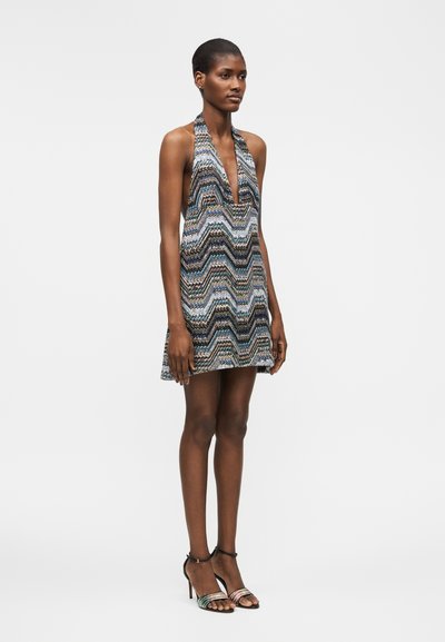 Γυναίκα στέκεται φορώντας μια αμάνικη, πολύχρωμη μίνι φόρεμα με ζιγκ-ζαγκ σχέδιο και ψηλοτάκουνα σανδάλια με λουράκια μπροστά σε λευκό φόντο.