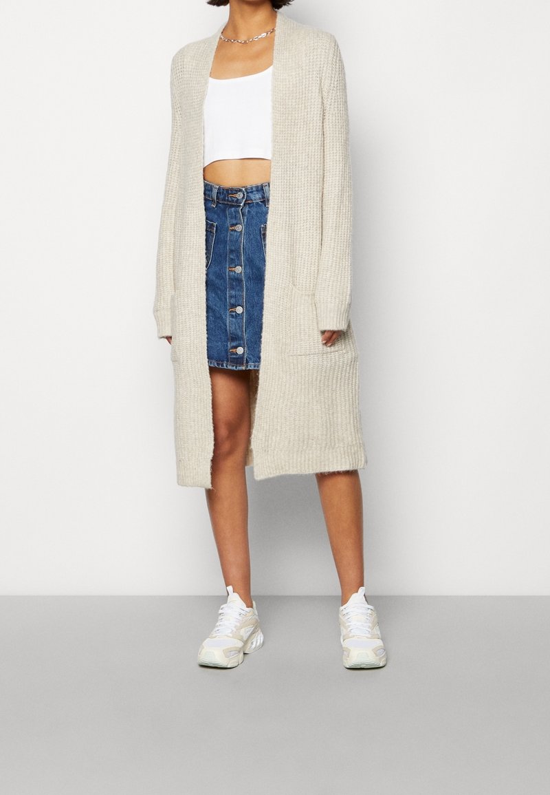 Langer beiger Strickcardigan mit offener Vorderseite, zwei Taschen, getragen über einem weißen Crop-Top und einem Jeansrock. Kombiniert mit weißen Turnschuhen.