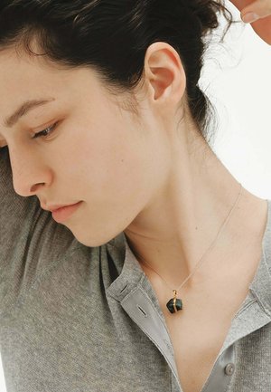 Joven mujer con camisa gris y un collar de plata con un colgante de piedra oscura, inclinando la cabeza hacia un lado con los ojos cerrados.