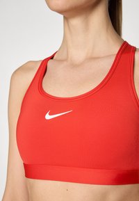 Rode sportbeha gemaakt van gladde, rekbare stof. Heeft een gebogen halslijn en een wit Nike swoosh-logo in het midden.