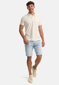 Cremfarbener Poloshirt mit kurzen Ärmeln, kombiniert mit hellblauen Jeansshorts. Mit aufgerollten Säumen und beigen Sneakers. Einfaches Design, lässige Passform.