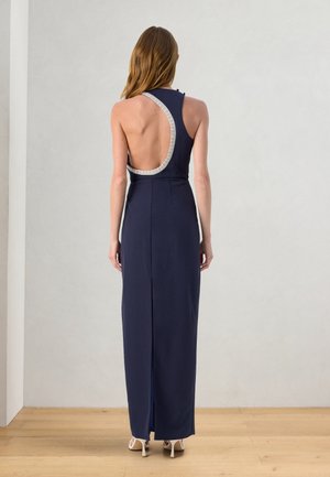 Donna in piedi con la schiena rivolta che indossa un abito lungo blu navy senza maniche con schiena aperta asimmetrica e dettaglio di bordo bianco.