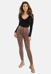 Donna con capelli lunghi e ondulati che indossa un top nero a maniche lunghe, leggings marroni a vita alta con motivo e cerniera frontale, e scarpe da ginnastica beige.