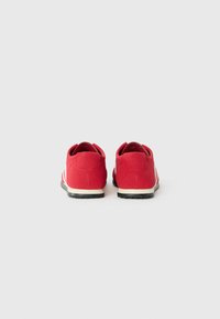 Czerwone buty sportowe z zamszowego materiału, zaokrąglonym noskiem, wyściółką wokół kostki i czarną gumową podeszwą. Posiadają białe paski ozdobne po bokach.