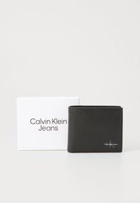 Black leather wallet with a smooth texture, featuring a logo on the lower right, displayed beside a white box with "Calvin Klein Jeans" printed on it.