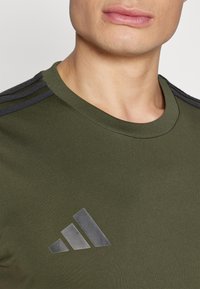 Olijfgroene sportshirt met ronde hals, voorzien van donkergrijze drie-strepen accenten op de schouders en een reflecterend logo op de borst.