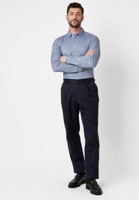 Man staat met gekruiste armen, gekleed in een blauw-wit gestreept shirt, donkere marineblauwe broeken en zwarte leren schoenen tegen een witte achtergrond.