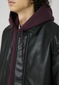 Veste en cuir noir avec un sweat à capuche bordeaux en dessous. Présente un col classique, une fermeture éclair et des cordons sur le sweat à capuche. Texture lisse.