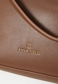 Borsa in pelle marrone con texture, caratterizzata da un design a forma curva nella parte superiore e dal logo dorato "ANNA FIELD". Il rivestimento liscio esalta l'aspetto complessivo.