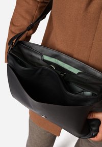 Sort læder messenger taske med en glat tekstur, der har en indvendig lynlås-lomme og grøn foring. Justerbar rem til bæring.