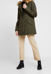 Parka vert olive avec capuche bordée de fourrure, dotée de boutons sur le devant, poches latérales et coupe droite. Associée à un pantalon beige et des bottes noires.