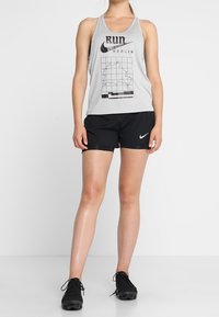 Canottiera senza maniche Nike grigia chiara con testo "RUN BERLIN" e grafica di una mappa, abbinata a pantaloni corti neri e scarpe da atletica nere.