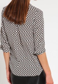 Kvinna med rakt brunt hår bär en långärmad blus med ett svart, vitt och persikofärgat geometriskt cirkelmönster.