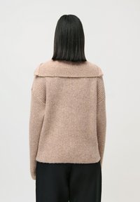 Person med rett, svart hår, ikledd en beige ribbestrikket genser og svarte bukser, sett fra bak mot en ensfarget bakgrunn.