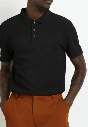 Mann trägt ein schwarzes kurzärmeliges Polohemd mit drei Knöpfen und rostfarbenen Hosen, Hände in den Taschen, blickt leicht nach links.