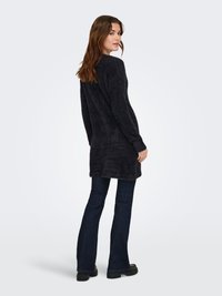 Pikkade pruunide juustega naine, kes kannab musta karvase tekstuuriga pikki kampsunit, tumedaid lehvivaid teksapükse ja musti paksu tallaga kingi, seistes veidi seljaga pööratult.