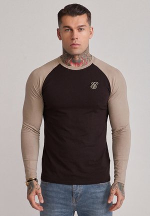 Mann med nakketatoveringer iført en svart og beige langermet skjorte og blå jeans, stående foran en ensfarget lys bakgrunn.