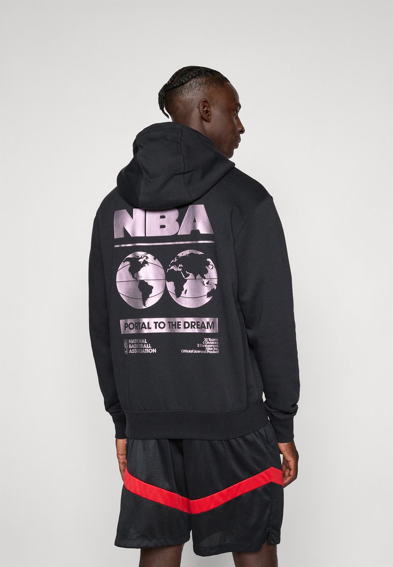 Mann med flettehår iført en svart NBA-hettegenser med lilla globusgrafikk og svarte basketballshorts med en rød stripe, sett fra siden.