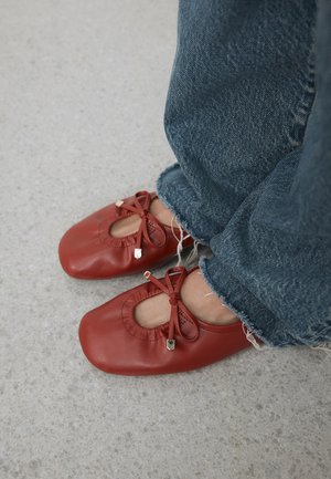 Person iført slidte blå jeans og røde læderballetflade sko med sløjfer, står på en tekstureret lysegrå gulv.