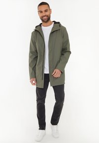 Veste imperméable vert olive avec capuche, fermeture éclair à l'avant et boutons-pression, associée à un jean noir et des chaussures blanches. Design simple, texture lisse.