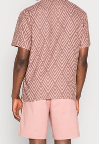 Man som bär en rosa geometriskt mönstrad kortärmad skjorta och enhetliga rosa shorts, stående med ryggen mot en neutral bakgrund.