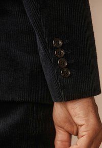 Dark corduroy blazer sleeve with a textured surface, featuring four black buttons and subtle stitching details.