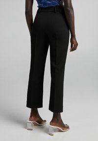 Kvinna klädd i svarta beskurna skräddarsydda byxor, blåmönstrad topp och genomskinliga klackar med öppen tå står på en grå golv.