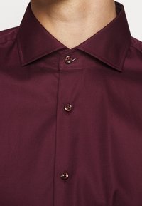 Gros plan d'une personne portant une chemise habillée bordeaux foncé boutonnée avec un col pointu.