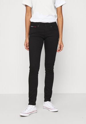 Personne portant un pantalon noir ajusté, une chemise blanche à manches courtes et des baskets blanches montantes, debout devant un fond blanc uni.