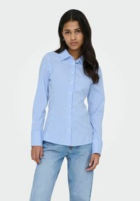 Chemise à rayures bleues et blanches avec boutons, manches longues, col pointu et larges poignets, associée à un jean en denim clair.