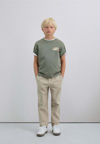 Camiseta de algodón verde con mangas enrolladas, que presenta un pequeño gráfico en el pecho, combinada con pantalones beiges y zapatillas blancas.