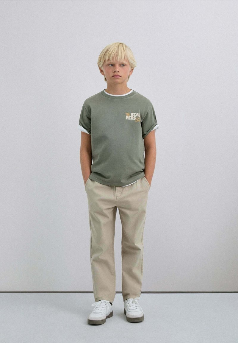 Camiseta de algodón verde con mangas enrolladas, que presenta un pequeño gráfico en el pecho, combinada con pantalones beiges y zapatillas blancas.