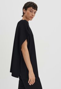 Haut noir style cape avec des manches larges, texture tricot douce, ourlet droit et fentes latérales pour le confort, associé à un pantalon sombre.