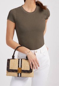 Sac à main en paille tissée avec des accents sombres à motifs, une fermeture à clip et une poignée courte, assorti à un haut côtelé olive et un pantalon blanc.