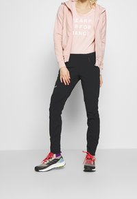 Ljusrosa zip-up hoodie, beige tröja med "PEAK PERFORMANCE" text, svarta byxor och mångfärgade sneakers med röda snören och detaljer.