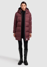 Bordeaux péřová bunda s kapucí, prošívaný design a přední zip. Nosená přes černé legíny a černé kozačky po kolena.