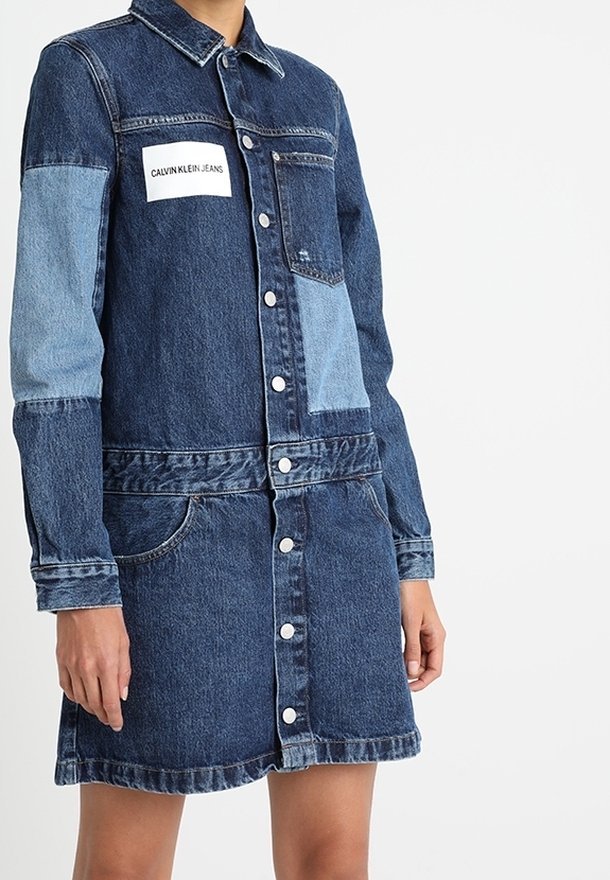 Denim dress with dark blue wash and light blue panel on one sleeve. Features button closure, two front pockets, and a branded patch.