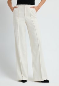Szerokie nogawki, kremowe spodnie z plisami z przodu i bocznymi kieszeniami, wykonane z gładkiego materiału. Stylizowane z czarnymi butami.