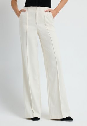 Szerokie nogawki, kremowe spodnie z plisami z przodu i bocznymi kieszeniami, wykonane z gładkiego materiału. Stylizowane z czarnymi butami.