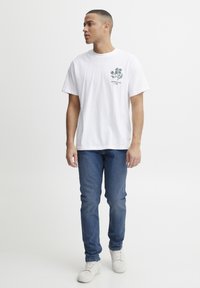 Camiseta blanca de algodón con un diseño floral verde en el lado izquierdo del pecho, combinada con jeans azules y zapatillas blancas.