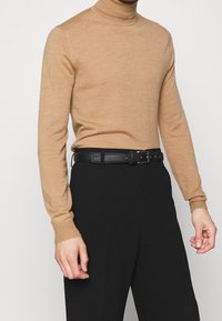Homme portant un pull à col roulé beige à manches longues rentré dans un pantalon noir taille haute avec une ceinture en cuir noire.