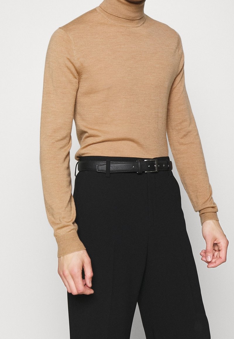 Homme portant un pull à col roulé beige à manches longues rentré dans un pantalon noir taille haute avec une ceinture en cuir noire.