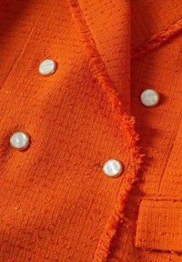 Oranžová texturovaná látka s třásněmi a čtyřmi kulatými bílými knoflíky se zlatými okraji na detailu oblečení.