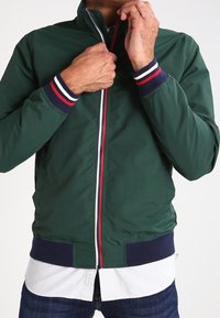 Muž zapínající tmavě zelenou bundu s manžetami a pasem s pruhy v námořnické modři, bílé a červené barvě, přes bílou košili a tmavé džíny.